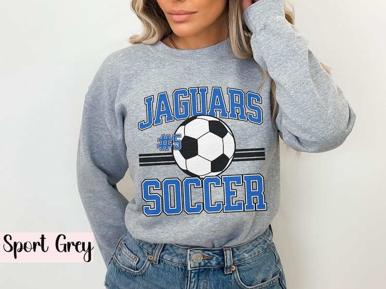 K&ouml;nnte beinhalten: Sportgraues Sweatshirt mit dem Text "JAGUARS SOCCER" in blauen Buchstaben und einer Fu&szlig;ballgrafik. Die Nummer "5" befindet sich &uuml;ber dem Fu&szlig;ball. Das Sweatshirt hat einen Rundhalsausschnitt.