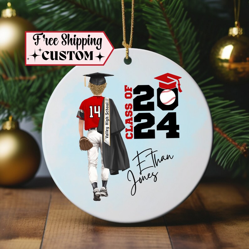 May include: A white ceramic ornament with a blue background. It features a cartoon illustration of a baseball player wearing a red and white uniform, a black graduation cap and gown, and holding a baseball. The text "Class of 2024" is in black and red, and the name "Ethan Jones" is written in cursive.