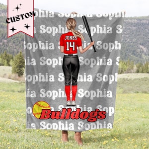 May include: Gray fleece blanket with a softball player illustration. The player is wearing a red jersey with the name "JONES" and the number "14" on the back. The blanket also has the text "Bulldogs" and the name "Sophia" repeated multiple times.