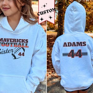 May include: White hooded sweatshirt with the text "MAVERICKS FOOTBALL Sister 44" on the front and "ADAMS 44" on the back. The sweatshirt has a front pocket and a hood.