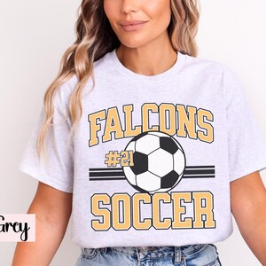 K&ouml;nnte beinhalten: Hellgraues T-Shirt mit einem Fu&szlig;ball-Grafik. Das Shirt zeigt das Wort "Falcons" &uuml;ber einem Fu&szlig;ball und das Wort "Soccer" darunter. Die Nummer "21" befindet sich &uuml;ber dem Fu&szlig;ball.