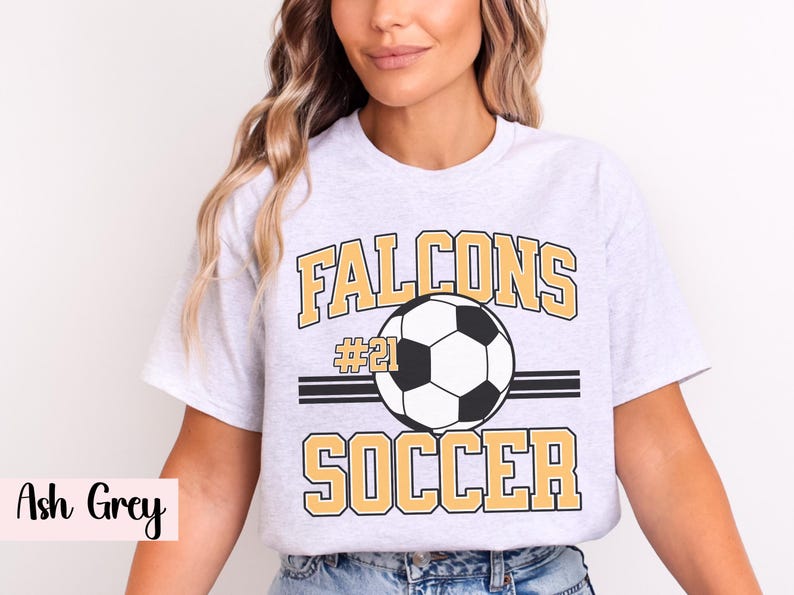 K&ouml;nnte beinhalten: Hellgraues T-Shirt mit einem Fu&szlig;ball-Grafik. Das Shirt zeigt das Wort "Falcons" &uuml;ber einem Fu&szlig;ball und das Wort "Soccer" darunter. Die Nummer "21" befindet sich &uuml;ber dem Fu&szlig;ball.