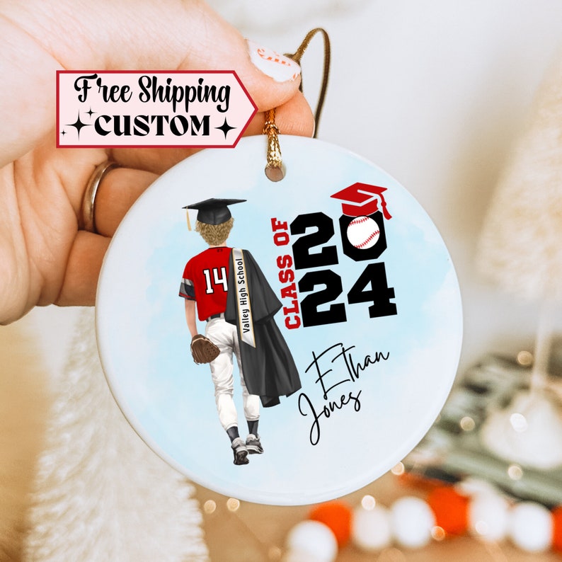 May include: A white ceramic ornament with a blue background featuring a cartoon illustration of a baseball player wearing a red and white uniform with the number 14 on the back. The player is wearing a graduation cap and gown. The text "CLASS OF 2024" is written in black with a red baseball on the top of the 2. The text "Ethan Jones" is written below the number 24. The text "Valley High School" is written on a red and white sash.