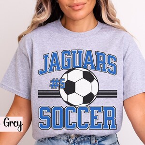 K&ouml;nnte beinhalten: Sportgraues T-Shirt mit einem Fu&szlig;ball-Grafikdesign. Das Design zeigt das Wort "Jaguars" in blauen Blockbuchstaben &uuml;ber einem Fu&szlig;ball und das Wort "Soccer" darunter. Die Nummer "5" ist ebenfalls enthalten.
