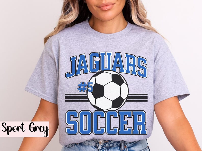 K&ouml;nnte beinhalten: Sportgraues T-Shirt mit einem Fu&szlig;ball-Grafikdesign. Das Design zeigt das Wort "Jaguars" in blauen Blockbuchstaben &uuml;ber einem Fu&szlig;ball und das Wort "Soccer" darunter. Die Nummer "5" ist ebenfalls enthalten.