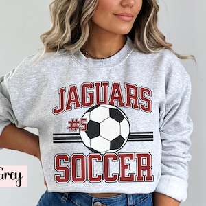 K&ouml;nnte beinhalten: Hellgraues Sweatshirt mit dem Text "JAGUARS SOCCER" in roter und wei&szlig;er Schrift, mit einer Fu&szlig;ballgrafik. Die Nummer "#5" befindet sich &uuml;ber dem Fu&szlig;ball. Das Sweatshirt ist ein Rundhals-Stil.