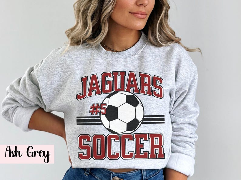K&ouml;nnte beinhalten: Hellgraues Sweatshirt mit dem Text "JAGUARS SOCCER" in roter und wei&szlig;er Schrift, mit einer Fu&szlig;ballgrafik. Die Nummer "#5" befindet sich &uuml;ber dem Fu&szlig;ball. Das Sweatshirt ist ein Rundhals-Stil.