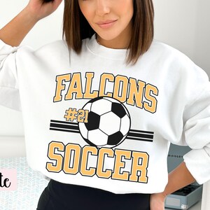 K&ouml;nnte beinhalten: Wei&szlig;es Sweatshirt mit dem Aufdruck "FALCONS SOCCER" in goldfarbenen Buchstaben und einer Fu&szlig;ballgrafik. Die Nummer "21" befindet sich &uuml;ber dem Fu&szlig;ball. Das Sweatshirt hat einen Rundhalsausschnitt.