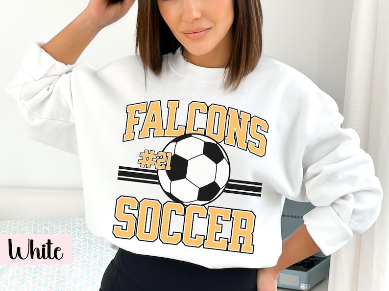 K&ouml;nnte beinhalten: Wei&szlig;es Sweatshirt mit dem Aufdruck "FALCONS SOCCER" in goldfarbenen Buchstaben und einer Fu&szlig;ballgrafik. Die Nummer "21" befindet sich &uuml;ber dem Fu&szlig;ball. Das Sweatshirt hat einen Rundhalsausschnitt.