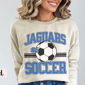 K&ouml;nnte beinhalten: Beigefarbenes Sweatshirt mit dem Wort "JAGUARS" in blauen Blockbuchstaben &uuml;ber einer Fu&szlig;ballgrafik. Unter dem Ball steht das Wort "SOCCER" in blauen Blockbuchstaben. Die Nummer "5" befindet sich &uuml;ber dem Fu&szlig;ball.