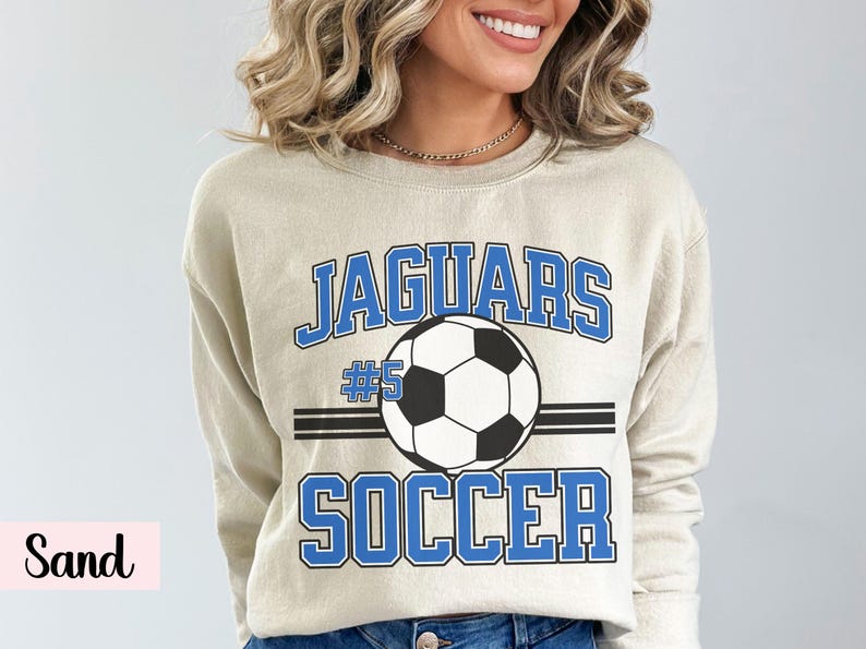 K&ouml;nnte beinhalten: Beigefarbenes Sweatshirt mit dem Wort "JAGUARS" in blauen Blockbuchstaben &uuml;ber einer Fu&szlig;ballgrafik. Unter dem Ball steht das Wort "SOCCER" in blauen Blockbuchstaben. Die Nummer "5" befindet sich &uuml;ber dem Fu&szlig;ball.