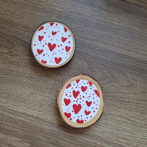 Puede incluir: Dos posavasos redondos de madera con fondo blanco y diseños de corazones rojos. Los posavasos tienen un borde de madera natural y están colocados sobre una superficie de madera. Los corazones son de diferentes tamaños y están dispersos por la superficie.