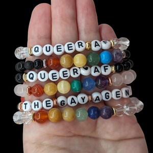 Peut inclure: Un ensemble de six bracelets avec des perles de pierres précieuses colorées et des perles de lettres blanches qui épellent "Queer AF" et "The Gay Agenda".