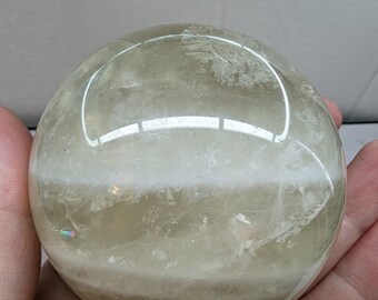 Op de afbeelding: Een doorschijnende, bolvormige kristallen bol met een lichtgele en witte kleurovergang. De kristallen bol wordt in een hand gehouden, waarbij het gladde, gepolijste oppervlak wordt getoond. De bol heeft een diameter van ongeveer 7.6 cm.