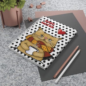 Puede incluir: Cuaderno de espiral con una ilustración de gato. El gato lleva una bufanda y sostiene una taza. La portada del cuaderno tiene un patrón de lunares blancos y negros con las palabras "Rose Notes" y un corazón rojo.