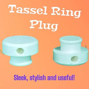 May include: Two white Tassel Ring Plugs on an orange background. The plugs are cylindrical with a hole in the center. Text on the image reads "Tassel Ring Plug" and "Sleek, stylish and useful!"