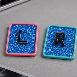 Op de afbeelding: Twee rechthoekige markers met afgeronde hoeken, één met een roze rand en de andere met een turquoise rand. Beide bevatten een met blauwe glitters gevuld midden met een verhoogde zwarte letter, een L en een R.