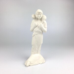 Peut inclure: Figurine en céramique blanche représentant une personne portant deux agneaux. La figurine porte des robes simples et se tient sur une base en forme de mouton. La sculpture a une finition mate et est placée sur un fond uni.