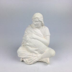 Peut inclure: Figurine en céramique blanche représentant une personne assise tenant un agneau. La personne est drapée d'un châle et porte une tunique simple. L'agneau est blotti dans les bras de la personne, créant une scène paisible. La figurine est non peinte.