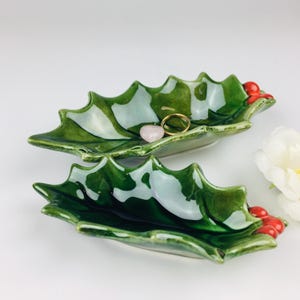 Peut inclure: Deux plats en céramique en forme de feuille, vert brillant avec des reflets blancs. Chaque plat a des accents de baies rouges et contient une bague dorée et une pierre rose. Les plats sont probablement pour bijoux ou petits objets.