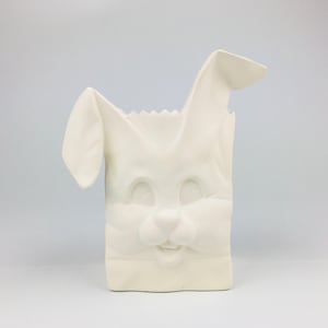 Peut inclure: Un récipient en céramique blanc en forme de lapin, avec un visage souriant et des oreilles dressées. Le récipient a une base rectangulaire et un bord supérieur en zigzag, ressemblant à un sac en papier. Les traits du lapin sont sculptés en relief.