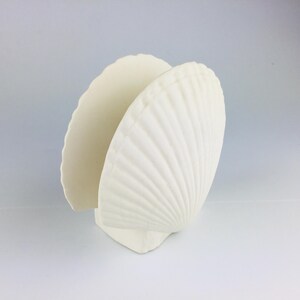 May include: A white seashell-shaped napkin holder. The holder is made of a solid, opaque material and features a detailed, ridged texture. The design is reminiscent of a scallop shell, perfect for coastal decor.