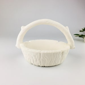 Peut inclure: Panier en céramique blanc cassé avec un extérieur texturé et une anse incurvée. La poignée est conçue pour ressembler à une branche d'arbre. Le panier est vide et repose sur une surface blanche.