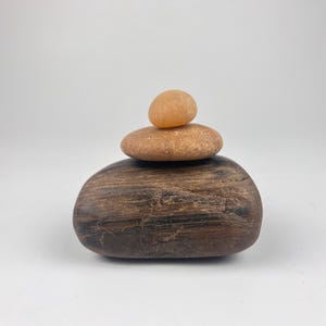May include: A stack of three stones in varying shades of brown and orange. The largest stone is a dark brown, oblong shape. A medium-sized, round, light brown stone sits atop it, with a small, orange stone at the top. The stones are arranged in a balanced, stacked formation.