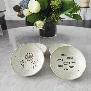 Könnte beinhalten: Vier kleine, gesprenkelte Keramikplatten mit unregelmäßigen Rändern. Zwei Teller zeigen schwarze Pusteblumen-Designs, während die anderen beiden einen Fischschwarm haben. Die Teller stehen auf einer reflektierenden Oberfläche, mit einer Blumenvase im Hintergrund.