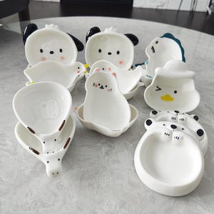 Puede incluir: Colección de platos de cerámica blanca con forma de animales, como un panda, un pato, una foca y un tigre. Cada plato tiene detalles pintados en negro, rosa y amarillo. Estos platos decorativos son ideales para servir pequeñas porciones.
