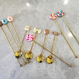 May include: A selection of gold-coloured spoons, each topped with a different candy design, such as donuts, lollipops, and a bow. The spoons are displayed on a light grey surface, highlighting their playful and decorative nature.