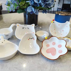 May include: A collection of white ceramic dishes in various animal shapes, including a bunny, cat, bear, duck, and chicken. One dish is shaped like a paw print with pink accents. The dishes are arranged on a light-colored surface.