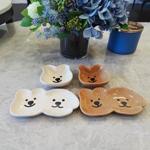 May include: Four ceramic bear-shaped dishes in white and brown, each with a black smiling face. Two dishes are single, and two are double. The dishes are on a marble-patterned surface, with a vase of flowers in the background.