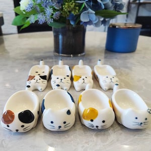 May include: A collection of white ceramic cat-shaped spoon rests and small dishes. Each piece features a different color pattern, including brown, black, and yellow spots, and blue accents. The cat-themed items are arranged on a light-colored surface.