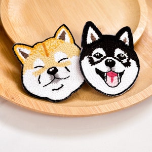 May include: Two embroidered patches featuring Shiba Inu dog faces. One patch has a smiling face with orange and white fur, while the other has a happy face with black and white fur and a pink tongue. The patches are on a wooden plate.