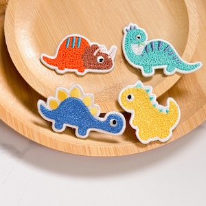 May include: Four embroidered iron-on patches featuring dinosaurs in various colors. The patches include a blue triceratops, a yellow stegosaurus, a red and orange triceratops, and a blue and green velociraptor.