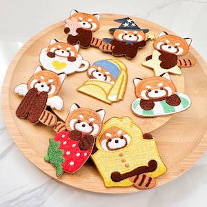 May include: A collection of embroidered patches featuring red pandas in various poses. The patches include a red panda holding a heart, wearing a witch hat, and inside a teacup. The patches are on a wooden tray.