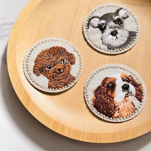 May include: Three embroidered patches featuring different dog breeds. The patches are beige with a white border and are on a wooden surface. The dogs are a brown poodle, a black and white schnauzer, and a brown and white cavalier king charles spaniel.