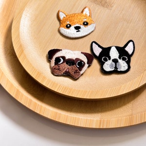 May include: Three embroidered iron-on patches featuring different dog breeds: a brown and white pug, a black and white French bulldog, and a brown and white Shiba Inu.