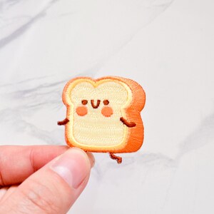 May include: Embroidered patch featuring a smiling slice of toast with arms and legs. The patch has a light tan center with an orange crust, rosy cheeks, and small brown details. The patch is held by a hand against a white background.