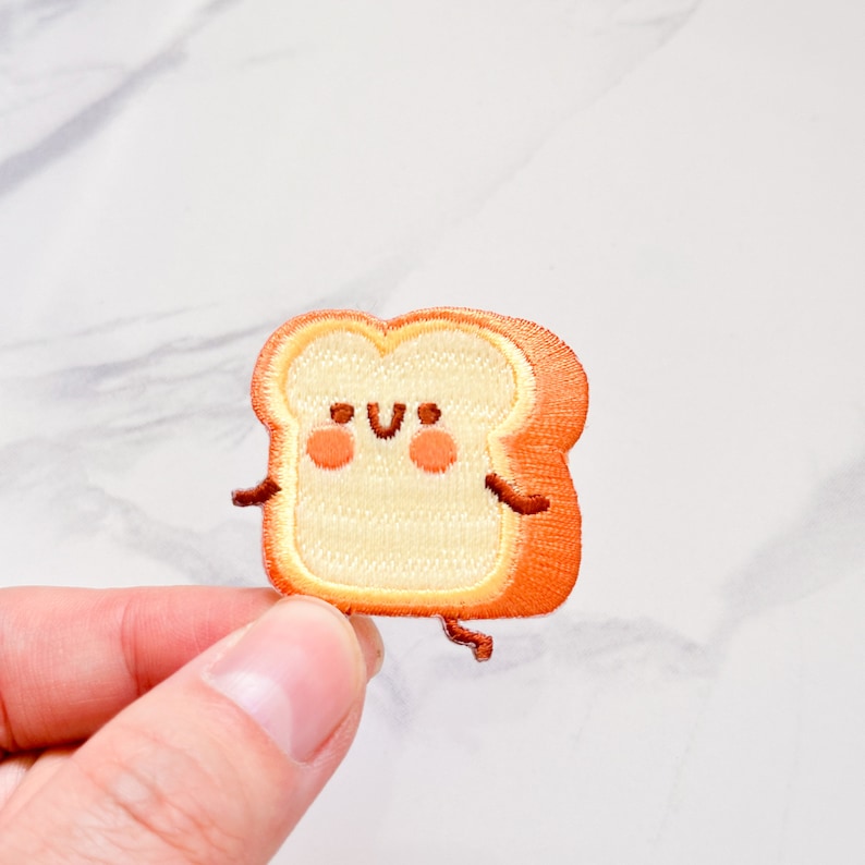 May include: Embroidered patch featuring a smiling slice of toast with arms and legs. The patch has a light tan center with an orange crust, rosy cheeks, and small brown details. The patch is held by a hand against a white background.