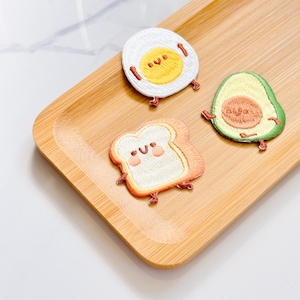 May include: Three embroidered patches: a fried egg, an avocado, and a slice of toast. The egg has a yellow yolk and white surrounding it. The avocado is green with a brown pit. The toast is tan with a white center. All three have smiling faces and small legs.
