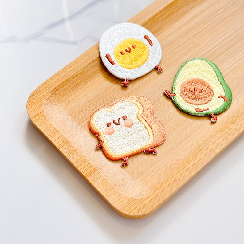 May include: Three embroidered patches: a fried egg, an avocado, and a slice of toast. The egg has a yellow yolk and white surrounding it. The avocado is green with a brown pit. The toast is tan with a white center. All three have smiling faces and small legs.