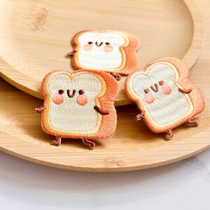 May include: Three embroidered patches shaped like slices of bread with smiling faces. The bread has a light tan center and orange crust, with small arms and legs. The patches are on a wooden plate.