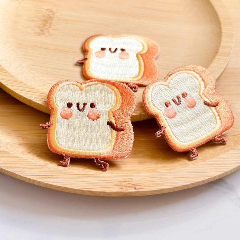 May include: Three embroidered patches shaped like slices of bread with smiling faces. The bread has a light tan center and orange crust, with small arms and legs. The patches are on a wooden plate.