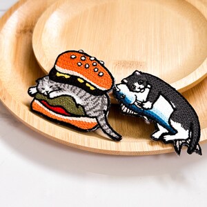 May include: Two embroidered iron-on patches featuring a cat eating a hamburger and a cat holding a fish. The patches are on a wooden surface.