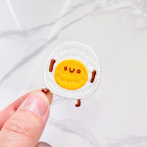 May include: Embroidered patch featuring a smiling fried egg design. The patch has a white background with a yellow yolk and brown details for the legs and arms. The egg has a happy face.