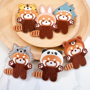 Peut inclure: Sept patchs brodés thermocollants représentant des pandas roux portant différents costumes d'animaux. Les patchs sont sur une surface en bois. Les costumes comprennent un tigre, un lapin, un requin, un panda, un ours et un loup.