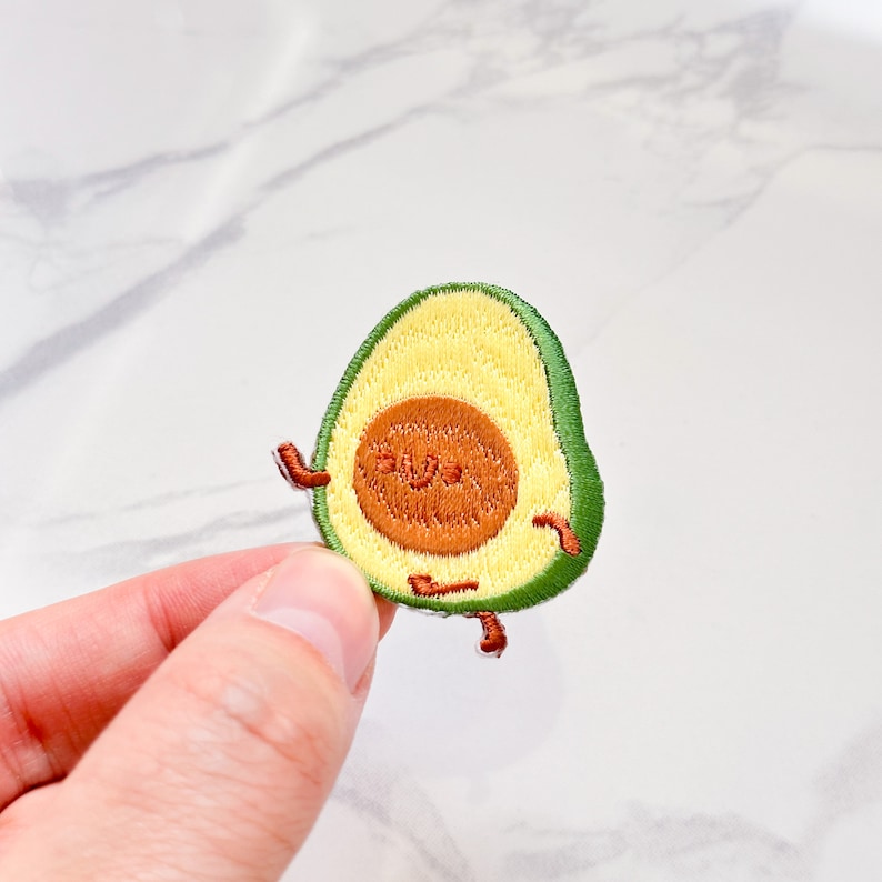 May include: Embroidered avocado patch with a smiling face, arms, and legs. The avocado has a green rind, yellow flesh, and a brown seed. The patch is held by a hand.