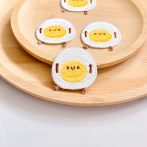 May include: Embroidered patches featuring a smiling fried egg design. The patches have a white egg white with a yellow yolk, brown arms and legs, and a smiling face. The patches are on a wooden tray.